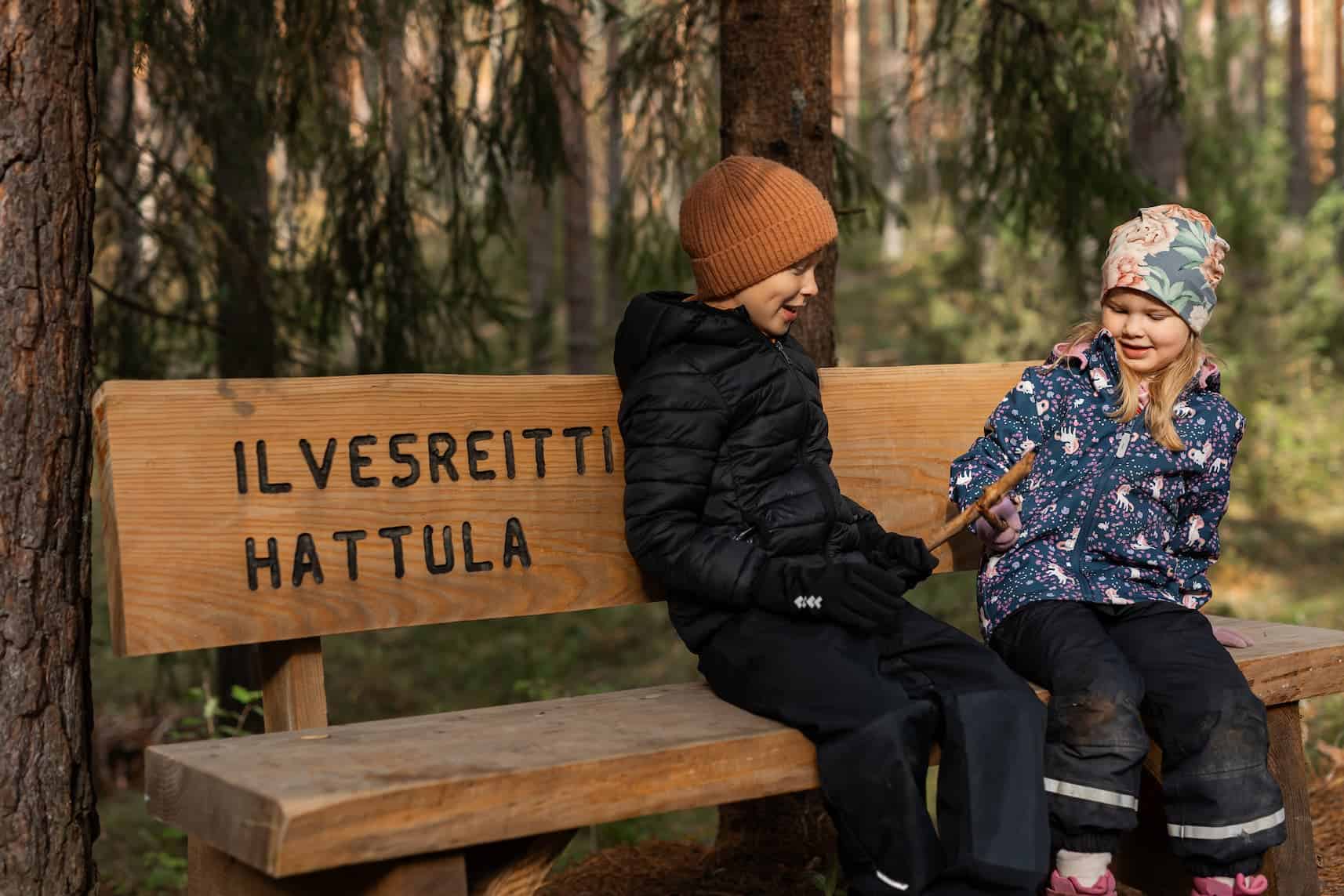 Kaksi lasta istuvat ulkona penkillä, johon on kirjailtu "Ilvesreitti, Hattula".